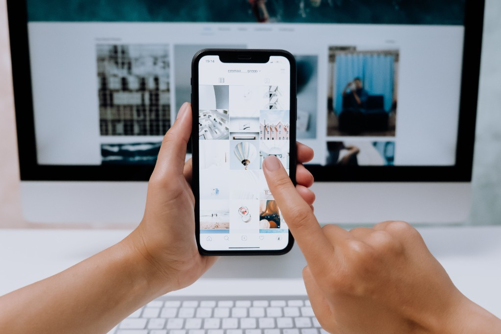 Hands holding a smartphone showing a vertical photo feed, with the same content on a desktop screen in the background